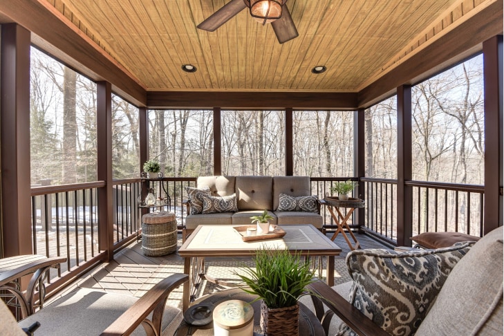 Screened-in porch with wood beam ceiling, woodgrain railing and framing between screens, and cushioned patio furniture. View of leafless trees outside.