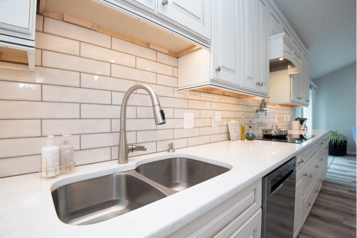 Remodeled kitchen with white cabinets, white stone countertop, stainless steel double sink, and white subway tile backsplash.